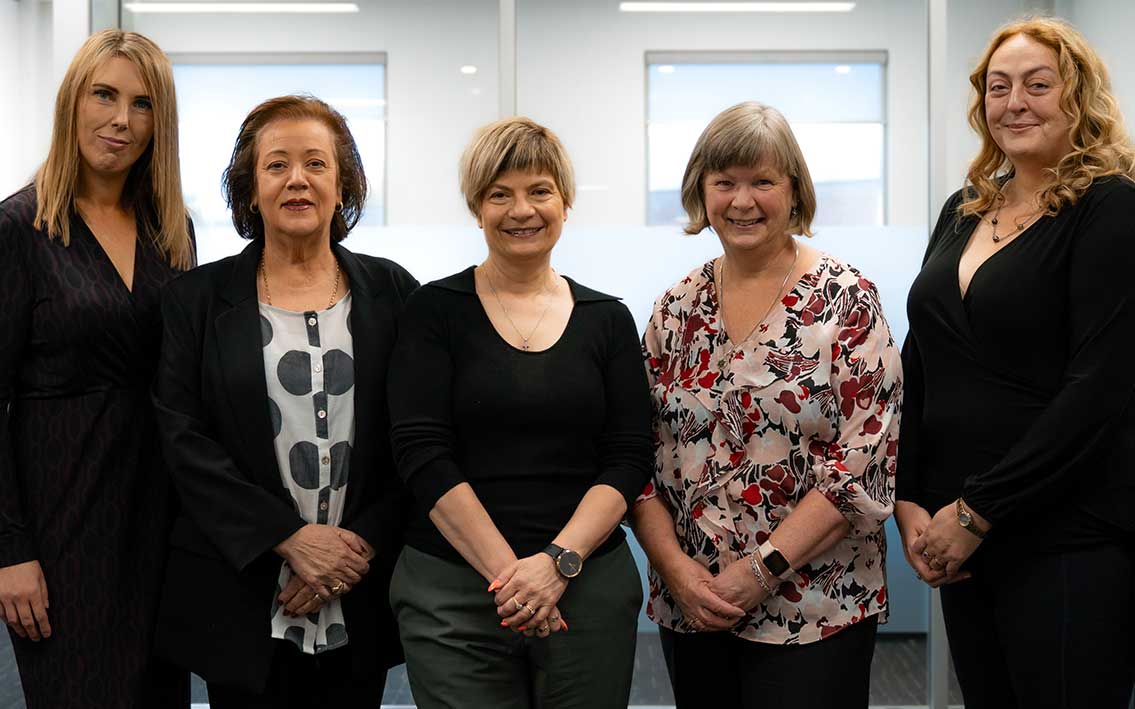 Five women standing indoors, all facing the camera and smiling. Five women standing indoors, all facing the camera and smiling.